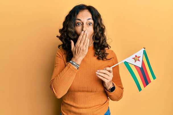 Middle age hispanic woman holding zimbabwe flag covering mouth with hand, shocked and afraid for mistake. surprised expression  - Foto, afbeelding
