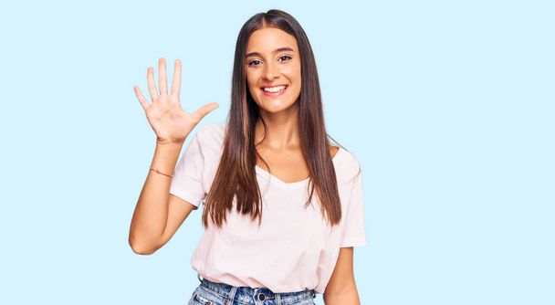 Mujer hispana joven vistiendo camiseta blanca casual mostrando y apuntando hacia arriba con los dedos número cinco mientras sonríe confiada y feliz.  - Foto, Imagen