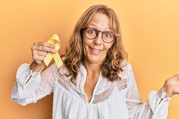 Mujer rubia de mediana edad sosteniendo la prevención del suicidio cinta amarilla celebrando el logro con sonrisa feliz y la expresión ganadora con la mano levantada  - Foto, imagen