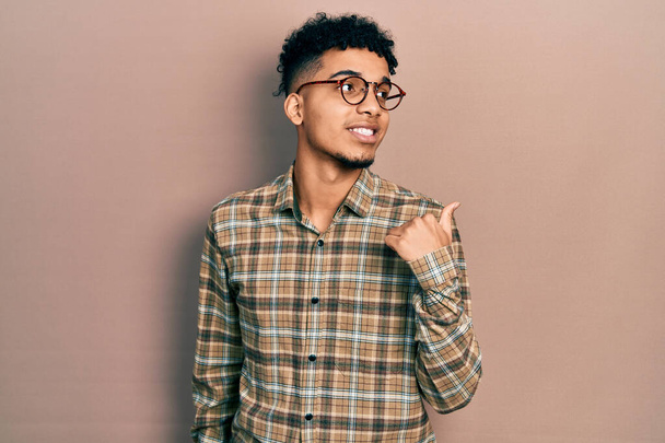 Νεαρός Αφροαμερικάνος που φοράει καθημερινά ρούχα και γυαλιά χαμογελώντας με χαρούμενο πρόσωπο και δείχνοντας στο πλάι με τον αντίχειρα ψηλά.  - Φωτογραφία, εικόνα