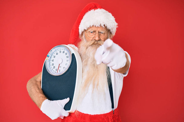 Старик на диете в традиционном костюме Санта Клауса показывает пальцем на камеру и на тебя, уверенный в себе жест выглядит серьезным  - Фото, изображение