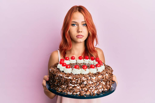 Νεαρή κοκκινομάλλα γιορτάζει τα γενέθλιά της με τούρτα χαλαρή με σοβαρή έκφραση στο πρόσωπο. απλή και φυσική κοιτάζοντας την κάμερα.  - Φωτογραφία, εικόνα