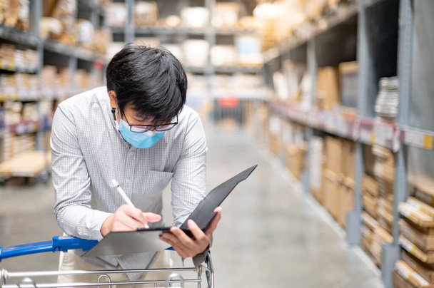 Asian man worker wearing face mask doing stocktaking of product in cardboard box on rack in warehouse by using clipboard. Physical inventory count and preventing the spread of COVID-19 (Coronavirus). - Photo, Image