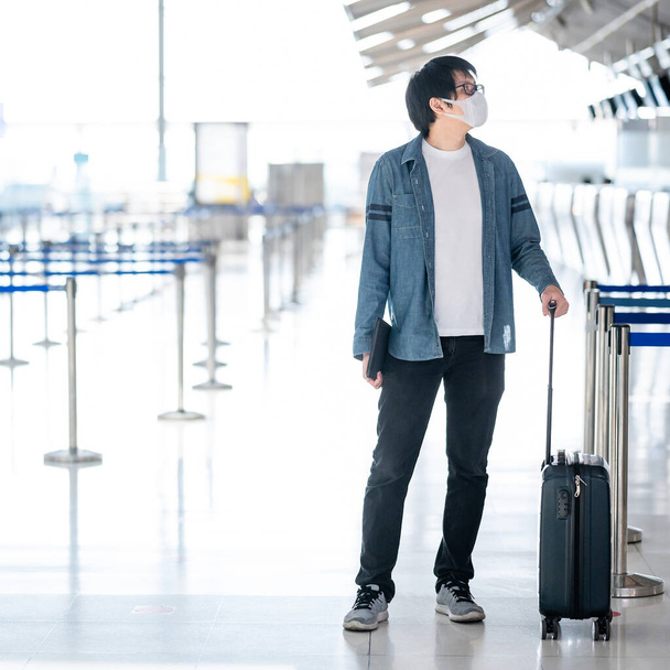 Ασιάτης τουρίστας φορώντας μάσκα προσώπου μεταφέρουν αποσκευές βαλίτσα στο check-in counter στο αεροδρόμιο τερματικό. Coronavirus (COVID-19) πρόληψη πανδημίας όταν ταξιδεύετε. Ευαισθητοποίηση και κοινωνική αποστασιοποίηση - Φωτογραφία, εικόνα