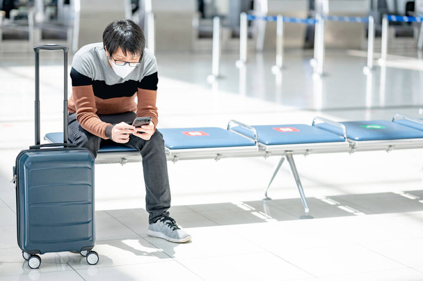 Asian man tourist with suitcase luggage wearing face mask using smartphone in airport terminal. Coronavirus (COVID-19) pandemic prevention when travel abroad. Health awareness and social distancing - Photo, Image
