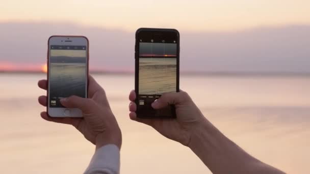 Unrecognizable man and woman holding smartphones in their hands taking pictures of sunset waterscape - Footage, Video
