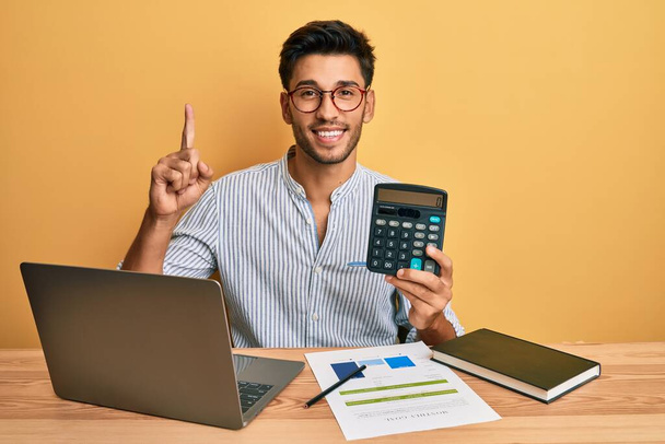 junger gutaussehender lateinischer Mann, der einen Taschenrechner an einen Computer anschließt und lächelt und eine erfolgreiche Geste zeigt - Foto, Bild