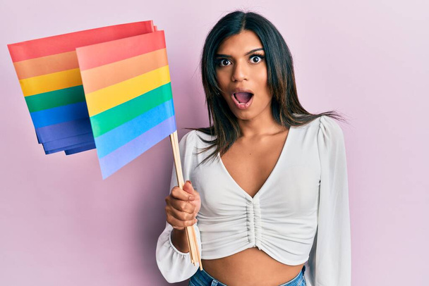 Jeune femme transsexuelle transsexuelle latine tenant le drapeau lgbt arc-en-ciel effrayé et étonné avec la bouche ouverte pour la surprise, visage incrédule  - Photo, image