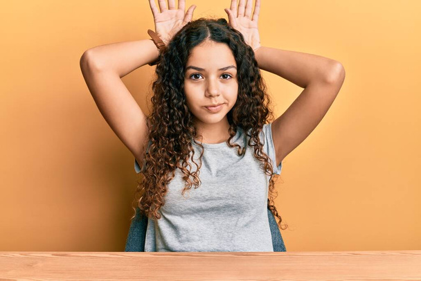 Teenager hispanic girl wearing casual clothes sitting on the table doing bunny ears gesture with hands palms looking cynical and skeptical. easter rabbit concept.  - Photo, Image