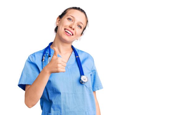 Young beautiful blonde woman wearing doctor uniform and stethoscope doing happy thumbs up gesture with hand. approving expression looking at the camera showing success.  - Photo, Image