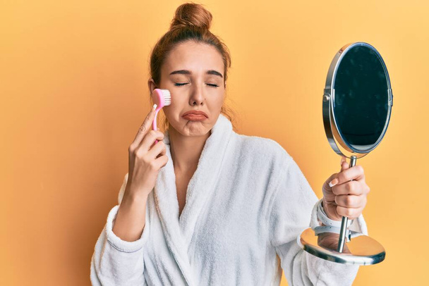 Young blonde woman wearing robe holding makeup remover brush depressed and worry for distress, crying angry and afraid. sad expression.  - Photo, Image