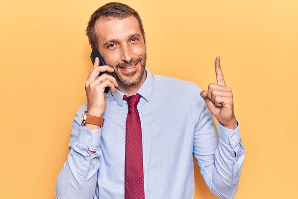 Junger gutaussehender Mann im Gespräch auf dem Smartphone lächelnd mit einer Idee oder Frage, Zeigefinger mit glücklichem Gesicht, Nummer eins  - Foto, Bild