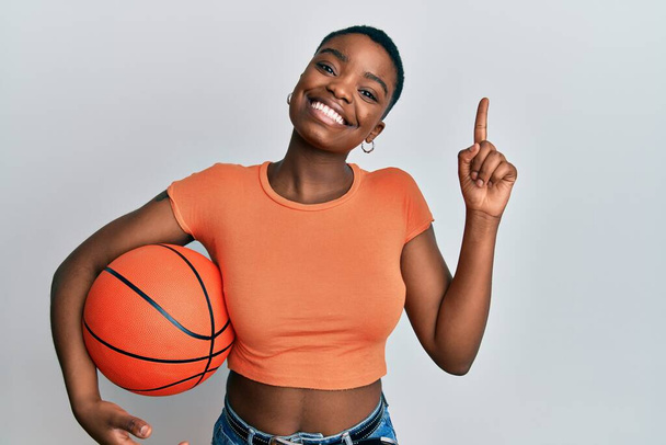 Jeune femme afro-américaine tenant ballon de basket souriant avec une idée ou une question pointant du doigt avec un visage heureux, numéro un  - Photo, image
