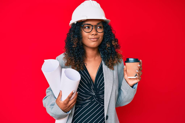 Young african american woman wearing architect hardhat holding blueprints and take away coffee smiling looking to the side and staring away thinking.  - Valokuva, kuva