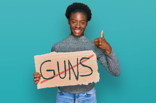 Young african american girl holding no guns warning banner smiling happy and positive, thumb up doing excellent and approval sign  - Foto, immagini