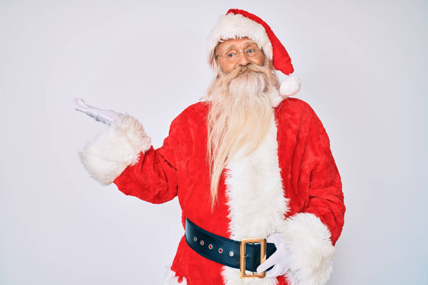 Alter älterer Mann mit grauen Haaren und langem Bart im Weihnachtsmann-Kostüm mit Hosenträgern, fröhlich lächelnd und mit der Handfläche in die Kamera zeigend.  - Foto, Bild