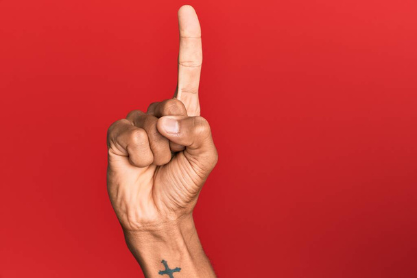 Mano del hombre hispano sobre fondo rojo aislado contando el número uno usando el dedo índice, mostrando idea y comprensión  - Foto, imagen