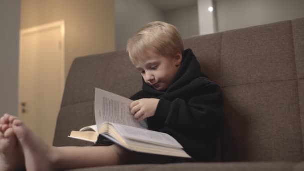 Netter kleiner Junge in schwarzem Kapuzenpulli sitzt auf der Couch und liest Bücher. Kleiner Junge blättert auf Couch - Filmmaterial, Video