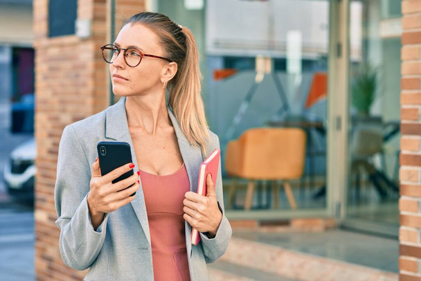 Young blonde businesswoman with serious expression using smartphone at the city. - 写真・画像