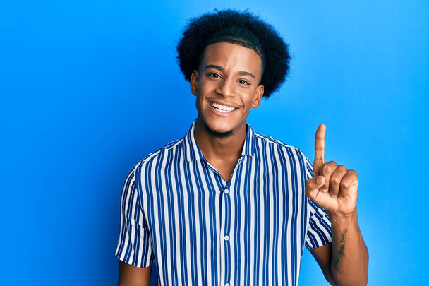 Afro-amerikanischer Mann mit Afro-Haaren, lässig gekleidet, lächelnd mit einer Idee oder Frage, erhobenen Zeigefinger mit glücklichem Gesicht, Nummer eins  - Foto, Bild