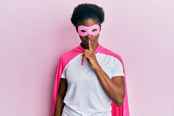 スーパーヒーローのマスクとケープの衣装を着た若いアフリカ系アメリカ人の女の子が唇に指で静かにするように求めています。沈黙と秘密の概念.  - 写真・画像