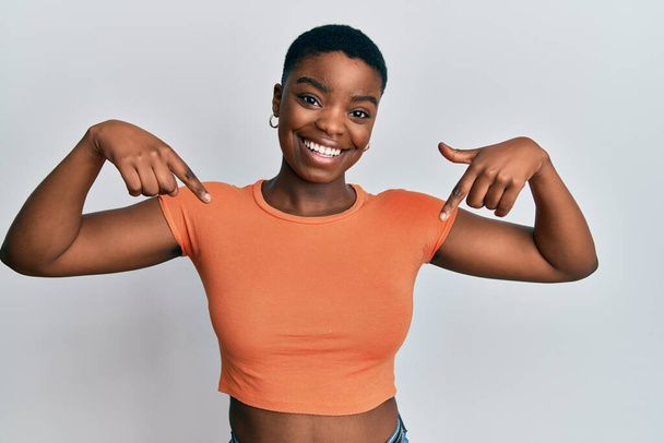 Fiatal afro-amerikai nő alkalmi narancssárga pólót visel, magabiztosan néz ki mosollyal az arcán, ujjaival büszkén és boldogan mutogatva magára..  - Fotó, kép