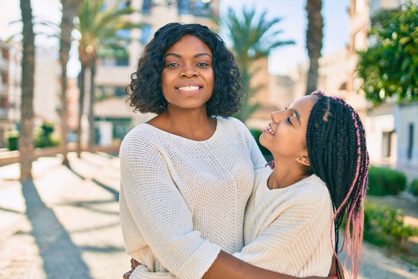 Hermosa madre afroamericana y su hija sonriendo feliz y abrazando. De pie con sonrisa en la cara de pie en el parque. - Foto, imagen