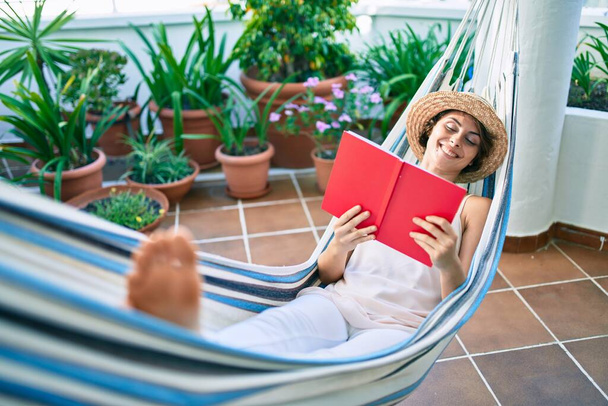 Fiatal, gyönyörű, fehér nő mosolyog, boldogan pihen egy függőágyon, könyvet olvas otthon. - Fotó, kép