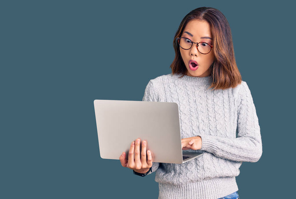 Jovem menina chinesa bonita usando óculos segurando laptop assustado e surpreso com a boca aberta para surpresa, rosto descrença  - Foto, Imagem
