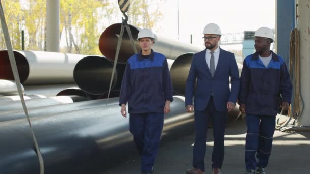  Медленная съемка мужчин и женщин, работающих на заводе в униформе и касках, показывающая стальные трубы мужчине-супервайзеру в костюме - Кадры, видео