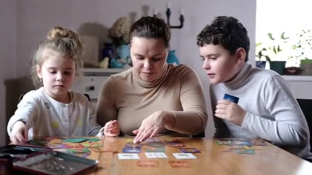 Bord educatieve spelletjes met het hele gezin thuis in een gezellig interieur. vrolijke jonge moeder en jongen en meisje spelen kaartspel aan tafel in de woonkamer. Gezamenlijke spelletjes van ouders en kinderen - Video