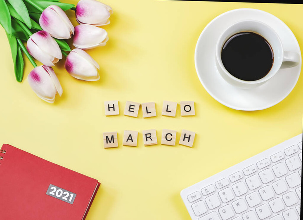 Top view or flat lay of woodeng letters HELLO MARCH, coffee cup, tulip flower bouquet, red diary2021 and computer keyboard on yellow background. Новый месяц и мотивация. - Фото, изображение