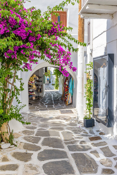 Tradycyjna uliczka rowerowa z wąską uliczką, wybieloną fasadą sklepów na zewnątrz kawiarni i kwitnącą bougainvillea na wyspie Naousa Paros, Grecja. - Zdjęcie, obraz
