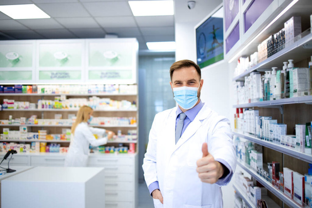 Портрет фармацевта в маске для лица и белом халате, показывающем большие пальцы в аптеке во время пандемии коронного вируса. - Фото, изображение