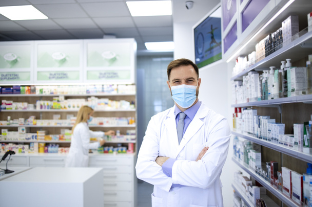 Portrait d'un pharmacien portant un masque facial et un manteau blanc debout dans un magasin de pharmacie pendant une pandémie du virus corona. - Photo, image