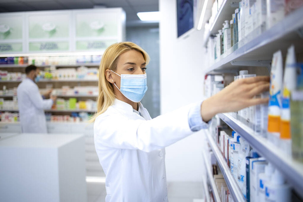 Vrouwelijke apotheker met gezichtsmasker en witte jas die in de apotheek werkt tijdens een corona-viruspandemie. - Foto, afbeelding