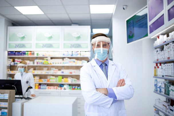 Portrait du pharmacien portant un écran facial et un manteau blanc debout dans un magasin de pharmacie pendant une pandémie de virus corona. - Photo, image