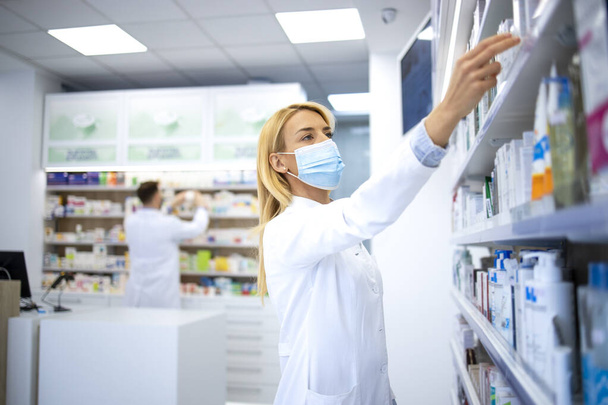 Vrouwelijke apotheker draagt gezichtsmasker en witte vacht houdt geneeskunde in apotheek op tijdens covid-19 virus pandemie. - Foto, afbeelding