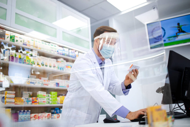 Фармацевт носит щит для лица и белое пальто, продавая витамины в аптеке во время пандемии коронного вируса. - Фото, изображение