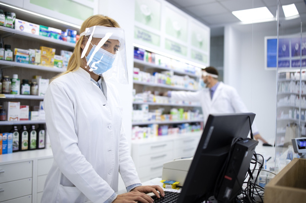 Kobieta blond farmaceuta kasjerka nosząca tarczę na twarz i biały płaszcz sprzedający witaminy w aptece podczas pandemii wirusa korony. - Zdjęcie, obraz
