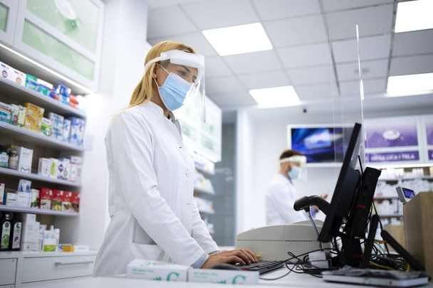 Γυναίκα ξανθιά φαρμακοποιός ταμίας φορώντας ασπίδα προσώπου και λευκό παλτό πώληση βιταμινών στο φαρμακείο κατά τη διάρκεια πανδημίας του ιού της κορώνας. - Φωτογραφία, εικόνα
