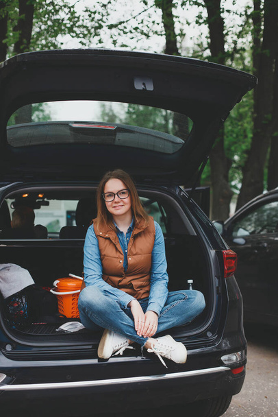 Mujer rubia feliz en el maletero abierto del vehículo todoterreno en el aparcamiento   - Foto, Imagen