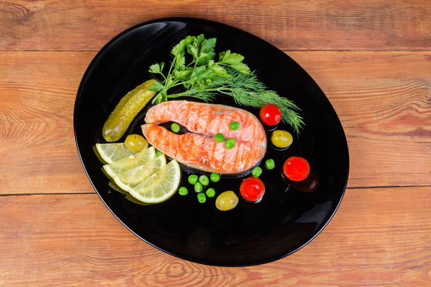 Filete a la parrilla de salmón, verduras enlatadas, rodajas de limón y verduras en plato negro sobre una mesa rústica de madera, vista superior - Foto, imagen