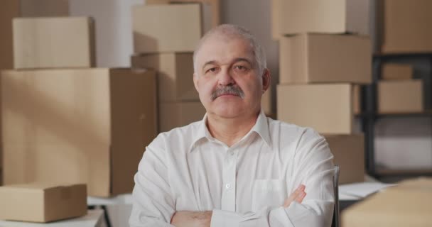 Portrait de propriétaire d'une petite entreprise d'un homme mûr avec moustache en chemise blanche plié ses bras regardant et souriant. Entrepôt avec marchandises dans des boîtes en carton. - Séquence, vidéo