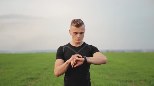 The athlete is standing on a field with green grass. He is choosing a workout on his watch. Running training. 4K - Footage, Video