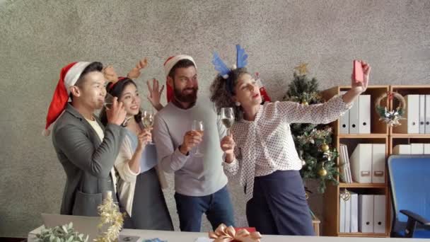 Fiatal nők és férfiak soknemzetiségű csapata ünnepli a karácsonyt az irodában, pezsgőt isznak, szelfit készítenek okostelefonon és boldogan mosolyognak - Felvétel, videó