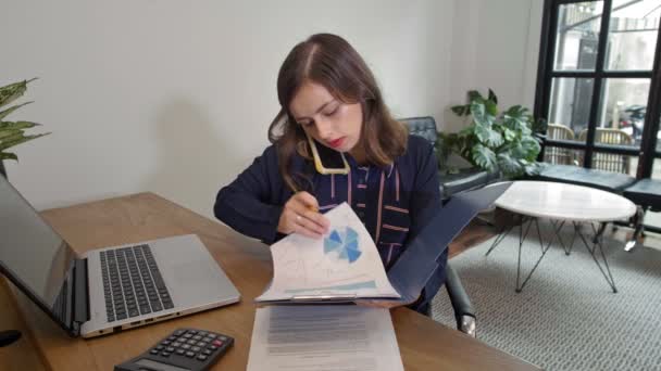 Medium shot of young Caucasian businesswoman sitting at desk, talking on phone about schemes in papers that she is holding in her hands and then typing something on laptop keyboard - Footage, Video