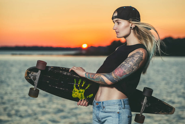 Ένα όμορφο, σέξι χίπστερ κορίτσι με τατουάζ στέκεται με μια σανίδα στο ηλιοβασίλεμα στη θάλασσα.. - Φωτογραφία, εικόνα