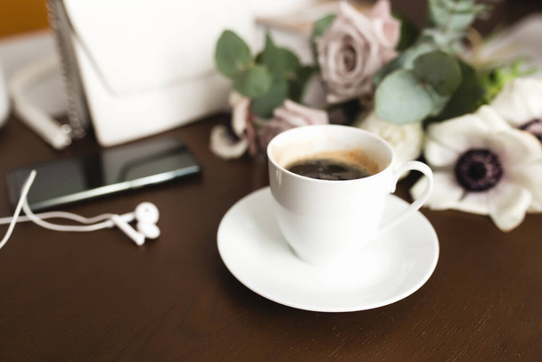 Una taza de café con un ramo de tierna anémona, ramas de eucalipto y rosas de color púrpura pastel, un bolso de mujer, auriculares y un teléfono en una mesa de madera marrón. Fondo borroso - Foto, imagen
