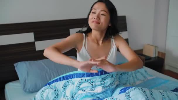 Genç, Asyalı, güzel bir kadın, battaniyenin altında yatakta oturuyor, uyandıktan sonra kendini geriyor ve mutlu bir şekilde gülümsüyor.  - Video, Çekim